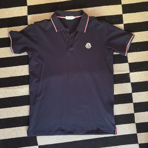 Mörkblå pikétröja från Moncler - Säljer en mörkblå pikétröja från Moncler med klassisk krage och knappar framtill. Tröjan har röda och vita ränder på krage och ärmslut samt Moncler-logga broderad på bröstet. Perfekt för en stilren och sportig look. Oanvänd priser kan diskuteras vid snabbt köp