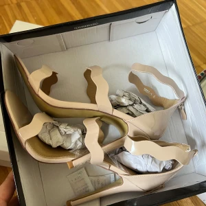 Beige klackade sandaler med vågig rem - Säljer ett par snygga beige sandaler med hög klack. Skorna har vågiga remmar i glansigt syntetmaterial och öppen tå. Helt oanvända. 