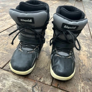 Svarta och grå snowboard boots från Liquid - Säljer ett par svarta och grå snowboard boots från Liquid med snörning och kardborreband. Skorna har quiltade detaljer på sidorna och en platt, mönstrad sula. Har några slitningar i tyget på några ställen men inga hål. 