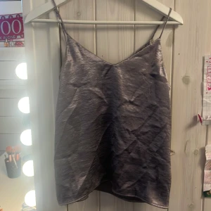 Grå satin linnetopp - Säljer en stilren grå linnetopp i satin med tunna axelband och v-ringad hals. Perfekt att styla till olika outfits för en trendig look.