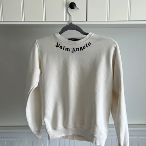 Vit sweatshirt från Palm Angels - Säljer en stilren vit sweatshirt från Palm Angels med svart logotyptryck både fram och bak vid halsen. Tröjan har rund halsringning och långa ärmar. Perfekt för dig som gillar streetwear och vill ha något enkelt men snyggt. Priset kan diskuteras 