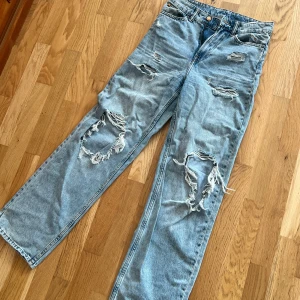 Ljusblå slitna straight jeans från H&M - Säljer ett par ljusblå jeans från H&M med straight passform och hög midja. Jeansen har flera slitningar och hål framtill och femficksmodell med knapp och dragkedja.