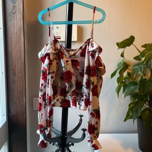 Vit blommig croppad linnetopp - Säljer en söt croppad linnetopp med röda rosor på vit botten. Toppen har tunna axelband, volangdetaljer och en luftig passform. Perfekt för varma dagar och ger en romantisk känsla till outfiten.