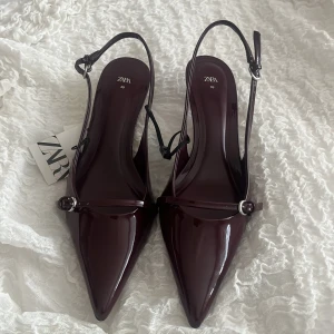 Vinröda lackade pumps från Zara i läder - Säljer ett par vinröda pumps från Zara med lackad finish och spetsig tå. Skorna har en smal rem med silverfärgat spänne och låg klack. Perfekta för dig som vill ha en elegant och trendig look. Aldrig använda. Lapp och säkerhetsband kvar. 