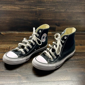 Svarta Converse Chuck Taylor All Star - Klassiska svarta Converse Chuck Taylor All Star high-top sneakers med vit sula och tå, samt vita snören. Skorna har rund tå och tydlig logotyp på sidan. Perfekta för en avslappnad och tidlös stil.