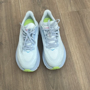 Hoka Clifton 9 löparskor i ljusblått - Säljer ett par Hoka Clifton 9 löparskor i ljusblått med detaljer i limegrönt och rosa. Skorna har snörning, rund tå och en tjock, dämpad sula för extra komfort. Perfekta för dig som gillar att springa eller promenera långt.