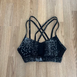 Svart sport-bh med vita prickar från Reebok - Snygg svart sport-bh från Reebok med vita prickar och flera smala korsade axelband. Perfekt för träning. Storlek XS. Pris 50kr