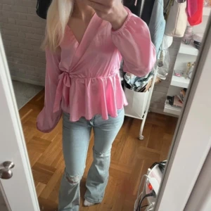 Rosa omlottblus med knyt  - Säljer en söt rosa omlottblus från Missay i glansigt material. Blusen har långa ärmar, volang nertill och knyt i midjan. Perfekt för dig som vill ha en feminin och trendig look.