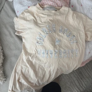 Beige t-shirt från Golden Goose - Snygg beige t-shirt från Golden Goose med trycket 'Golden Goose University' framtill i vitt. Klassisk rund halsringning och korta ärmar. Perfekt för en avslappnad och trendig stil.