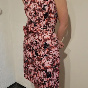 Blommig kortklänning från Lindex, stl 38 - Säljer en snygg blommig klänning från Lindex i rosa, rött och svart. Klänningen är ärmlös, har en markerad midja med bälte och en feminin passform. Perfekt för dig som vill sticka ut med ett färgstarkt mönster!