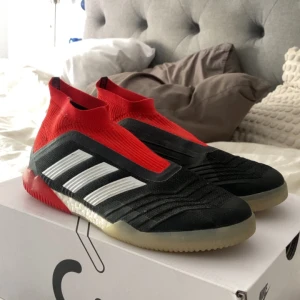 Adidas Predator 18+ in fotbollsskor - Säljer ett par Adidas Predator 18+ fotbollsskor i svart, rött och vitt. Skorna har en strumpliknande passform utan snörning, med tydliga vita ränder på sidan och röd häl. Sulan är ljus med röda detaljer undertill. Gjorda för inomhus men går ute med, kartan följer även med
