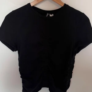 Svart croppad t-shirt från H&M - En svart croppad t-shirt från H&M med korta ärmar och rynkade detaljer framtill som sitter tajt och visar kurvor fint. Perfekt till jeans eller kjol för en enkel och stilren look. 