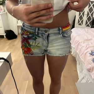Low waist jeans shorts - Säljer ett par ljusblå jeansshorts med färgglatt blommigt mönster på ena sidan framtill. Shortsen har klassisk femficksdesign och detaljer i olika färger vid linningen.