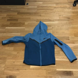 Blå hoodiejacka från Nike - Säljer en blå hoodiejacka från Nike med dragkedja och huva. Jackan har två olika blå nyanser och svarta detaljer längs dragkedjan och på ärmarna. Perfekt för dig som gillar sportig stil och vill ha något bekvämt och snyggt. (Junior storlek 147-158) väldigt sällsynt säljs inte längre
