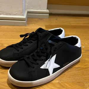 Svarta sneakers från Golden Goose med vit stjärna - Säljer ett par svarta sneakers från Golden Goose med vit stjärndetalj på sidan och vit häl. Skorna har svarta skosnören och vit sula. Perfekta för dig som gillar stilrena och trendiga sneakers.