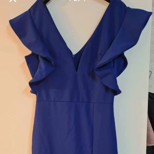 Marinblå klänning med volangdetaljer - Säljer en marinblå klänning med v-ringning och stora volangdetaljer vid axlarna. Klänningen har en elegant, figurnära passform och slits baktill för extra rörelsefrihet. Perfekt för dig som vill sticka ut med stil.