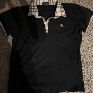 Burrbery Piké - Säljer en svart t-shirt från Burberry med klassisk beige, röd och svart rutigt krage. T-shirten har korta ärmar och liten broderad Burberry-logga på bröstet. Perfekt för dig som gillar stilrena plagg med lyxig touch.Storlek är XL i barn.