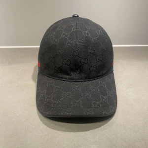 Svart gucci canvas keps - Svart keps från Gucci med diskret GG-monogram över hela ytan. Kepsen har en böjd skärm och justerbar rem baktill. Sidorna pryds av Guccis klassiska röd-gröna band. Perfekt accessoar för en stilren look.