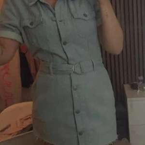 Kort jeansklänning med bälte - Säljer en snygg kort jeansklänning med knappar framtill och tillhörande bälte i midjan. Klänningen har korta ärmar med uppvik och två bröstfickor. Perfekt för en trendig och avslappnad look.