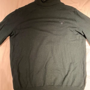 Mörkgrön polotröja från GANT - Säljer en klassisk mörkgrön polotröja från GANT med diskret broderad logga på bröstet. Tröjan är långärmad och tillverkad i mjukt material, perfekt för kyligare dagar. Enkel och stilren design som passar till mycket.