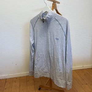 Grå ziptröja från BOSS - Säljer en stilren grå ziptröja från BOSS med hög krage och dragkedja hela vägen. Tröjan har två fickor framtill och diskret BOSS-logga på bröstet. Perfekt för en avslappnad och sportig look. Storlek L men sitter som M. 
