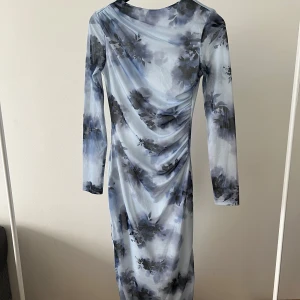 Blommig långklänning i blå och grå toner - Säljer en långklänning med blommigt mönster i olika nyanser av blått och grått. Klänningen har lång ärm och en draperad effekt längs sidorna. Perfekt för dig som gillar en elegant och stilren look.