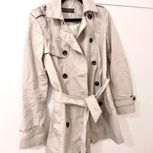 Beige trenchcoat med bälte - Stilren beige trenchcoat från Zara med klassisk dubbelknäppning och bälte i midjan. Jackan har långa ärmar, axelklaffar och knappar vid ärmslut. Perfekt för övergångssäsonger och ger en tidlös look.