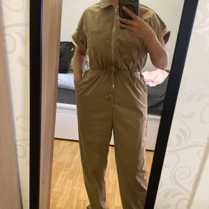 Beige jumpsuit med dragkedja och snörning i midjan - Säljer en beige jumpsuit med korta ärmar, dragkedja framtill och snörning i midjan. Modellen har raka ben och fickor på sidorna. Perfekt för en avslappnad och trendig look.
