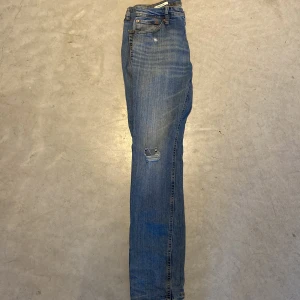 Blå slitna jeans med raka ben - Säljer ett par klassiska blå jeans med raka ben och slitningar på båda knäna. Jeansen har normal passform och är tillverkade i denim med synliga sömmar. Perfekta för en avslappnad stil.