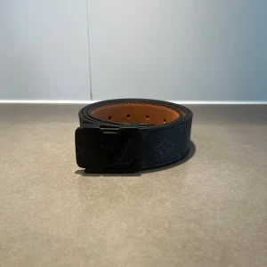 Svart LV bälte  - Säljer ett svart bälte från Louis Vuitton med klassiskt LV-monogram på både spännet och lädret. Bältet har en stilren design med svart utsida och brun insida. Perfekt accessoar för att lyfta din outfit.