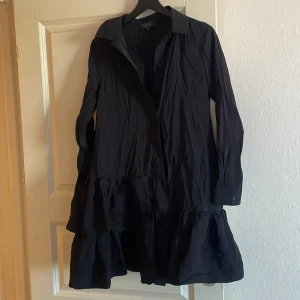 Svart skjortklänning från COS - Snygg svart skjortklänning från COS med krage och långa ärmar. Klänningen har en volangdetalj vid nederkanten som ger en fin touch. Perfekt för en stilren look.