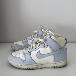 Nike Dunk High i ljusblått och vitt - Snygga Nike Dunk High sneakers i en fräsch kombination av ljusblått och vitt. Skorna har en klassisk hög design med snörning och en bekväm sula. Perfekta för en stilren look.