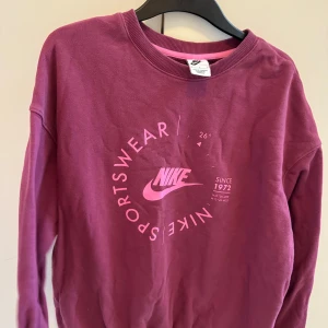 Vinröd sweatshirt från Nike - Säljer en vinröd sweatshirt från Nike med tryck i rosa på framsidan. Tröjan har långa ärmar och en rund halsringning. Perfekt för en sportig look eller avslappnade dagar.