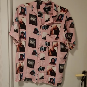 Rosa kortärmad skjorta med The Notorious B.I.G. motiv - Säljer en unik rosa skjorta från H&M med tryck av The Notorious B.I.G. Skjortan har korta ärmar och knappar framtill. Perfekt för dig som vill sticka ut med en cool och retro stil.