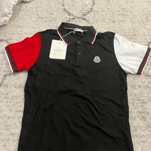 Svart pikétröja från Moncler - Snygg svart pikétröja från Moncler med kontrasterande röda och vita ärmar. Tröjan har en klassisk krage med knappar och Monclers logotyp på bröstet. Perfekt för en stilren look. Fraktas samma dag  passar M också 