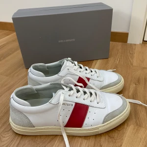  Axel Arigato sneakers - Snygga vita sneakers från Axel Arigato med en röd detalj på sidan. Skorna har snörning och en rund tå. Perfekta för en stilren look. Med  skoförpackning ingår även en grå påse med axel arigato märket. Priset kan diskuteras.