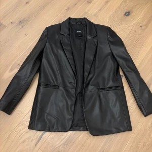 Svart skinnkavaj från Pull&bear - Snygg svart skinnkavaj från Pull&bear med klassisk design. Kavajen har en stilren look med långa ärmar och två fickor framtill. Perfekt för en elegant och modern stil.