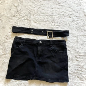 Svart jeanskjol med bälte - Snygg svart jeanskjol med medföljande bälte. Kjolen har en klassisk femficksdesign och stängs med knapp och dragkedja framtill. Perfekt för en cool och avslappnad stil.