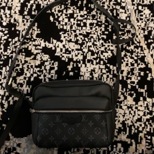 Svart axelväska med monogram - Snygg svart axelväska med monogrammönster och justerbar axelrem. Väskan har en dragkedja upptill och en extra ficka framtill med dragkedja. Perfekt för att bära dina dagliga nödvändigheter med stil.