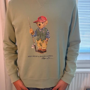 Grön sweatshirt med Polo Bear från Ralph Lauren - Säljer en grön sweatshirt från Ralph Lauren med det ikoniska Polo Bear-motivet på framsidan. Tröjan har långa ärmar och en rund halsringning. Perfekt för en avslappnad stil.