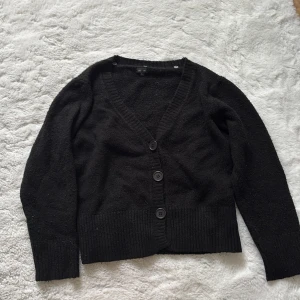 Svart kofta från Ze-Ze - Säljer en klassisk svart kofta från Ze-Ze med v-ringning och knappar framtill. Koftan har långa ärmar och ribbade detaljer vid ärmslut och nederkant. Perfekt för lager-på-lager-stil eller som ett enkelt basplagg i garderoben.