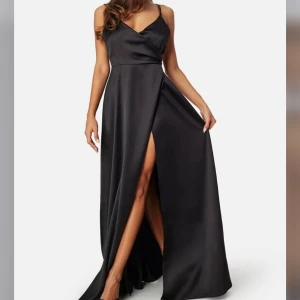 Svart långklänning med slits - Elegant svart långklänning i satin med smala axelband och v-ringad design. Klänningen har en stilren slits framtill och en öppen rygg med knytning. Perfekt för en glamorös kväll. Använts en gång.