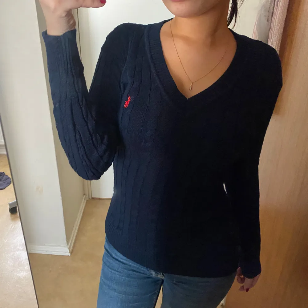 Säljer en stilren mörkblå stickad tröja från Ralph Lauren med v-ringning och ett diskret rött logomärke på bröstet. Tröjan har långa ärmar och en klassisk kabelstickad design. Perfekt för en avslappnad men ändå elegant look.. Neuletakit & Villapaidat.