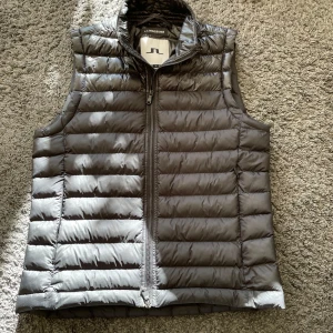 Svart dunväst från J.Lindeberg - Snygg svart dunväst från J.Lindeberg med dragkedja framtill. . Västen har en stilren design med horisontella quiltade paneler.