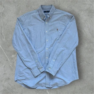 Polo Ralph Lauren skjorta Ljusblå - Säljer en klassisk ljusblå skjorta från Ralph Lauren med broderad logotyp på bröstet. Skjortan har långa ärmar och knappar framtill. Perfekt för en stilren look.