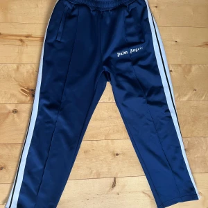 Blå träningsbyxor från Palm Angels - Snygga blå träningsbyxor från Palm Angels med vita ränder längs sidorna. Byxorna har en elastisk midja och sidofickor. Perfekta för en sportig look eller avslappnad stil. Storlek L