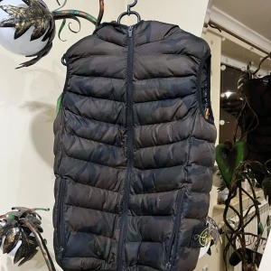 Camouflage dunväst från Stone Island - Säljer en cool dunväst från Stone Island i ett snyggt camouflagemönster. Västen har en dragkedja framtill och är perfekt för att hålla värmen. Den har också en orange detalj på insidan som ger en extra touch.Det ni ser på bilden är det ni får😁 ett plagg som är köpt i bulk så jag vet lila lite som ni, men jag har kollat så gott jag kan på alla möjliga sajter och det jag kan kolla stämmer överens med originalet väldigt bra tröja raka o fina sömmar, kvalitets tyg inledigt min åsikt 😁