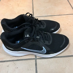Svarta Nike löparskor - Säljer ett par svarta Nike Quest löparskor med vit sula och ljusblå detaljer. Skorna har snörning och är designade för löpning. Perfekta för den som söker komfort och stil under träningspassen.
