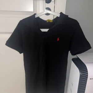 Svart kortärmad pikétröja från Ralph Lauren - Säljer en klassisk svart pikétröja från Ralph Lauren med korta ärmar och den ikoniska röda loggan på bröstet. Perfekt för en stilren och avslappnad look.