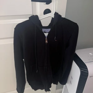 Svart hoodie från Tommy Hilfiger - Säljer en stilren svart hoodie från Tommy Hilfiger med dragkedja och justerbar huva. Perfekt för en avslappnad look. Den har en liten logga på bröstet och är i storlek S.